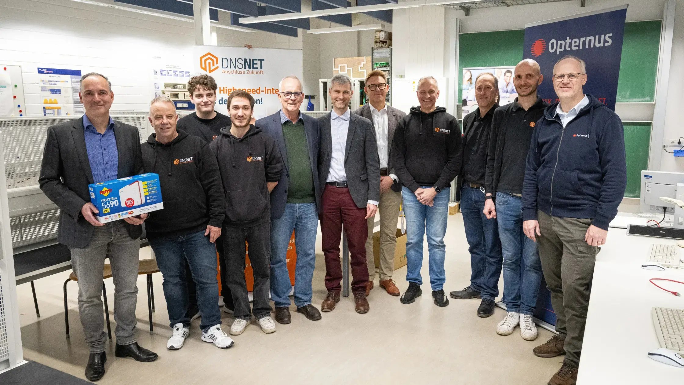 Gruppenfoto im Glasfaserlabor des OSZ IMT in Berlin: v. l. n. r. Martin Göhle (FRITZ!), Uwe Postel (DNS:NET), Jeremy Golze (DNS:NET), Jeremy Roger (DNS:NET), Michael Salner (OSZ IMT), Thorsten Basel (OSZ IMT), Volker Dahms (Schulleiter OSZ IMT), Volker Rau (DNS:NET), Andreas Görne (DNS:NET), Abel Enghy (DNS:NET) und Thomas Timm (Opternus) stehen gemeinsam vor Bannern von DNS:NET und Opternus; links hält Martin Göhle eine FRITZ!Box 5690.