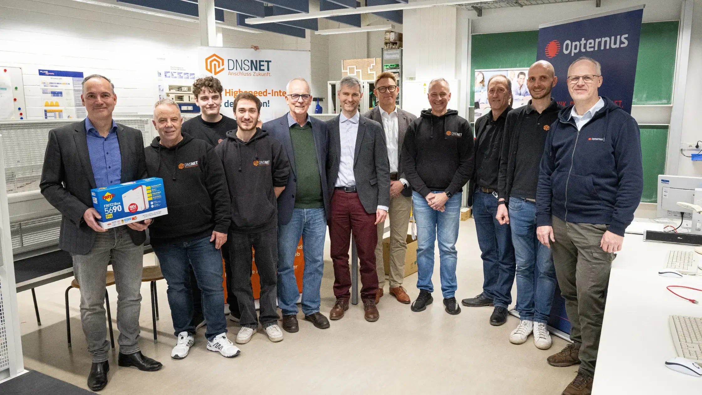 Gruppenfoto im Glasfaserlabor des OSZ IMT: Mitarbeitende von DNS:NET, Fritz! und Opternus sowie Lernende stehen gemeinsam vor DNS:NET- und Opternus-Roll-ups; links hält ein Mann eine FRITZ!Box 5690 in der Hand.